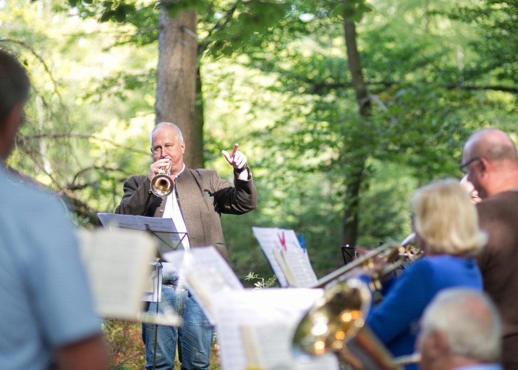 Mehrere Blasmusiker spielen zusammen. Vorne steht ein Trompeter und dirigiert mit einer Hand. Vor ihnen stehen Notenständer mit Liedblättern. Im Hintergrund Wald.
