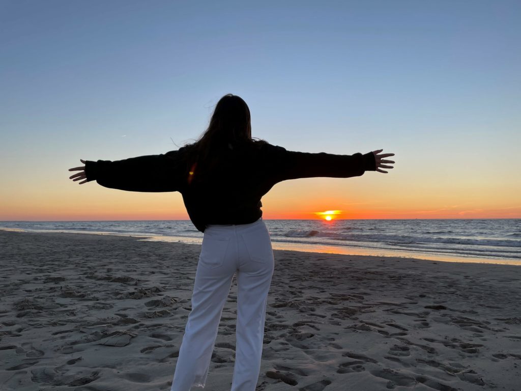 Eine Person steht mit ausgebreiteten Armen am Strand und schaut auf den Sonnenuntergang über dem Meer